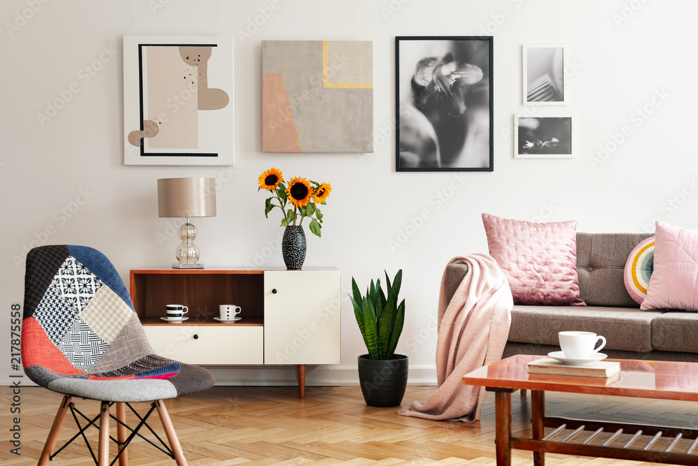 Obraz premium Modern chair and sunflowers on cabinet in flat interior with posters and pink pillows on sofa. Real photo