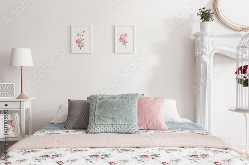 Pastel pillows on bed in feminine white bedroom interior with posters and lamp on cabinet. Real photo