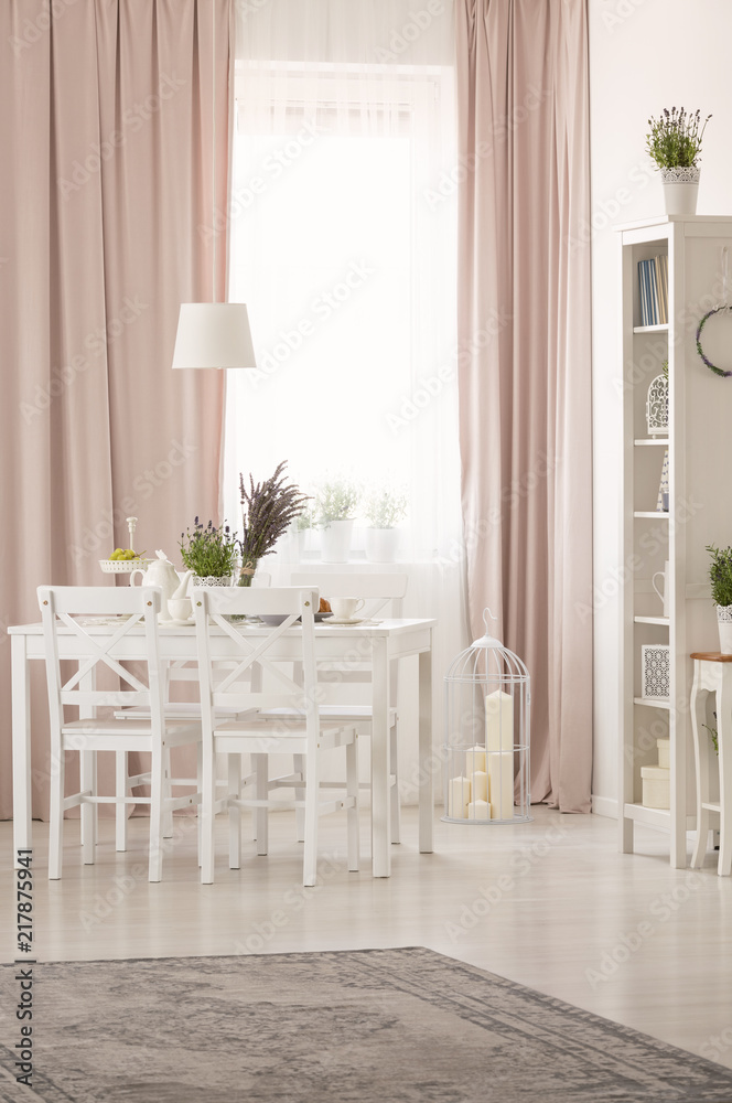 White chairs at table with plants in pastel pink dining room interior with rug and drapes. Real photo White chairs at table with plants in pastel pink dining room interior with rug and drapes. Real photo