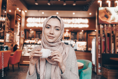 nice Arab girl with a headscarf on her head dines in a cozy cafe, drinks fragrant tea