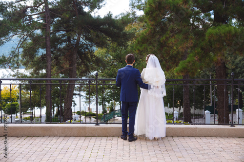 View from the back, newlyweds standing on the terrace, embracing looking at huge old trees, river and blue sky. The groom tenderly embraces the bride's waist.