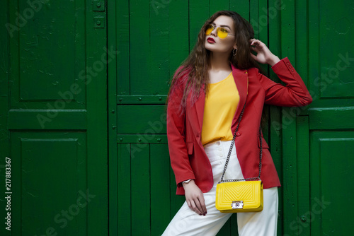 Outdoor fashion portrait of young beautiful woman wearing blazer, yellow sunglasses, blouse, white trousers, holding small quilted bag, posing near the green door. copy,empty space for text