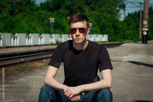 Portrait serious handsome young man, wearing casual clothing and sunglasses sitting on a step at railway station