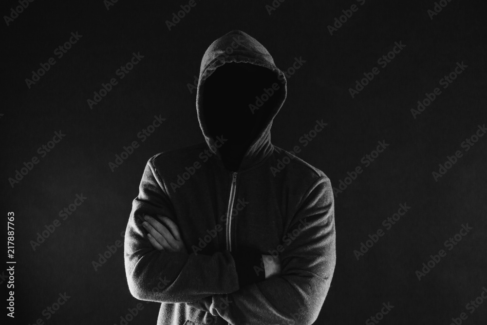 Anonymous and faceless man under hoodie with arms crossed isolated over dark background - incognito and mysterious criminal on internet activities concept. Anonymous and faceless man under hoodie with arms crossed isolated over dark background - incognito and mysterious criminal on internet activities concept.