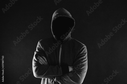 Anonymous and faceless man under hoodie with arms crossed isolated over dark background - incognito and mysterious criminal on internet activities concept.