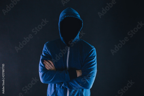 Anonymous and faceless man under hoodie with arms crossed isolated over dark background - incognito and mysterious criminal on internet activities concept.