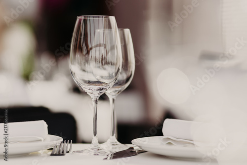 Empty glasses stand near plates and forks on banquet table decorated with white tablewear. Festively served table against blurred background in restaurant