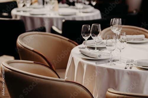 Horizontal shot of served round table with white tableclothes, wineglasses, plates, forks and knives, two armchairs near. Cozy atmosphere in luxury restaurant.