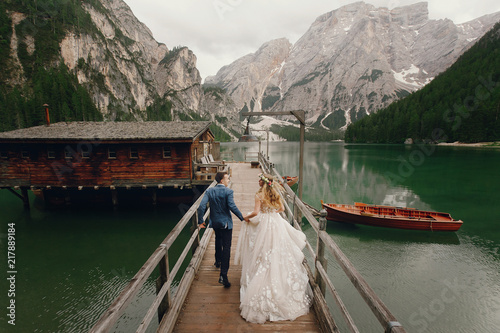Bride and groom hold each other hands walking along the bridge over the lake in Italian Dolomites photo