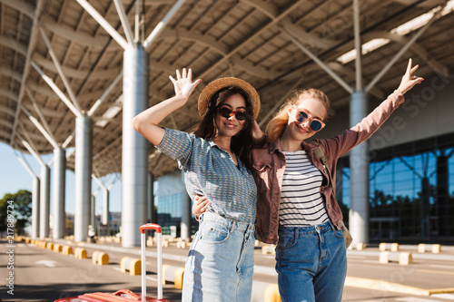 Two smiling girls in sunglasses joyfully looking in camera while raising hands up with airport on background