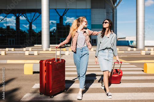 Beautiful stylish girls in sunglasses happily walking along pedestrian strip with suitcases and airport on background photo