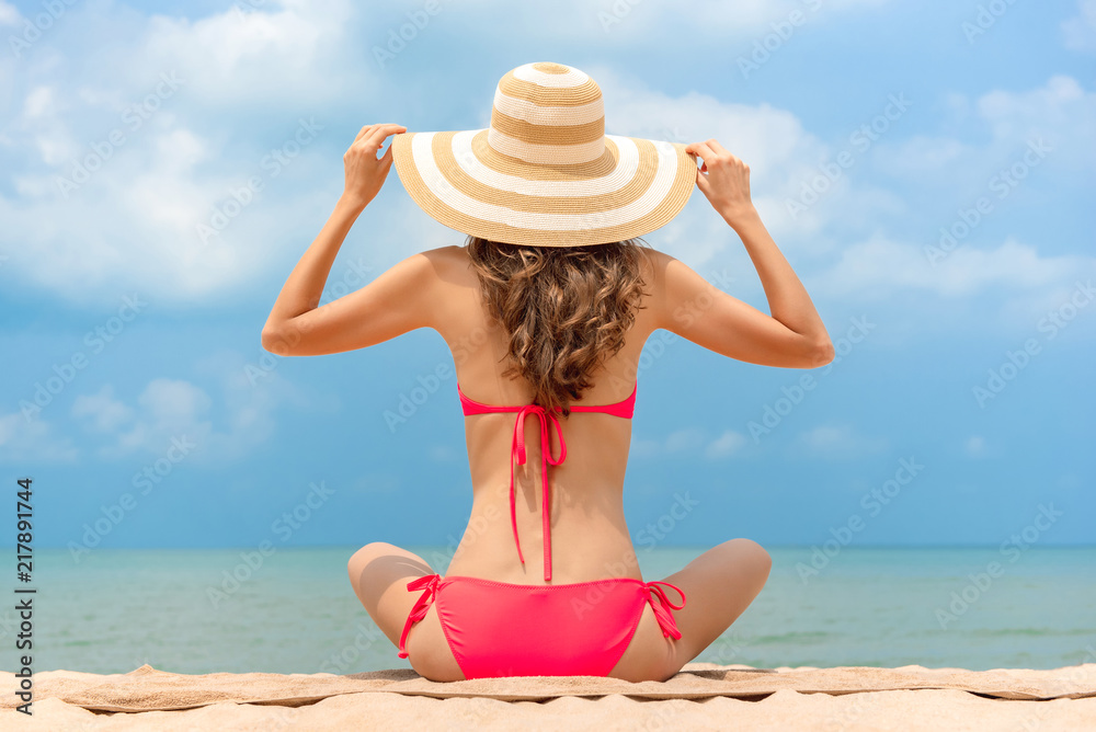 Woman in bikini with sun hat sitting at the beach in summer