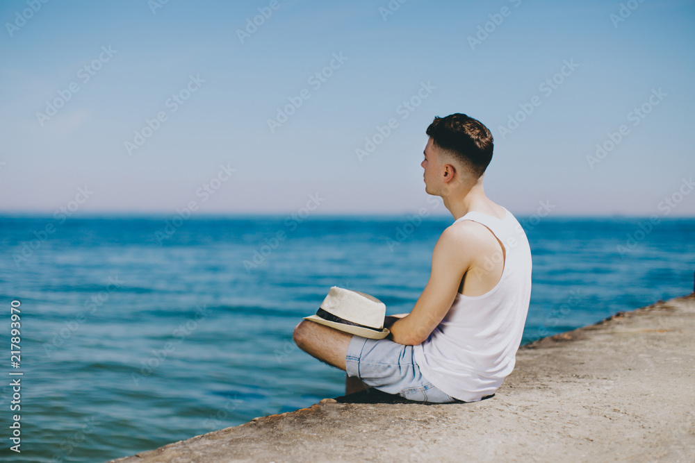 A young guy in a white T-shirt is sitting on the pier. He enjoys a beautiful view and thinks about future travels. What could be better than a holiday on the seaside.