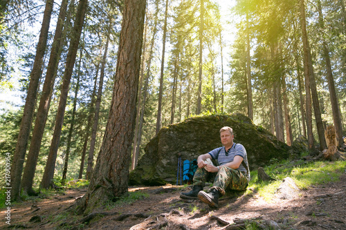 The tourist is resting near a large stone in the forest. A man sits on the ground during a holiday on a hike.