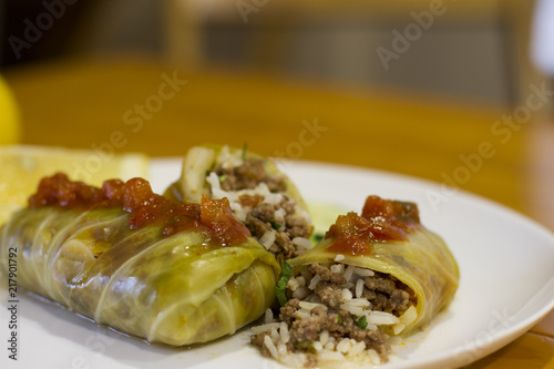 Cigar of cabbage served on white plate accompanied by slices of cucumber, sicilian lemon and tomato on yellow wooden table