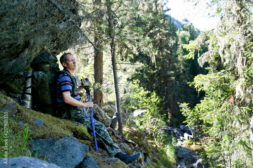 The tourist is resting under a large stone. A man with a backpack sits under a rock during a hike through the mountains and forest.