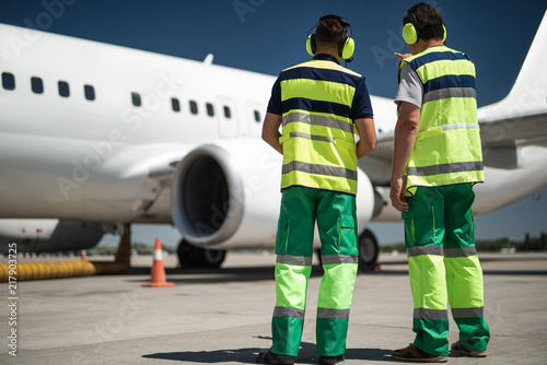 Let me show you. Back view of aviation personnel observing passenger aircraft. Man in headphones pointing at jet and talking with colleague