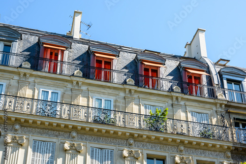 View from down the street of a typical parisian, opulent-looking building of haussmannian style with cut stone facade, carved ornaments, balcony and orange awnings on the top floor windows.