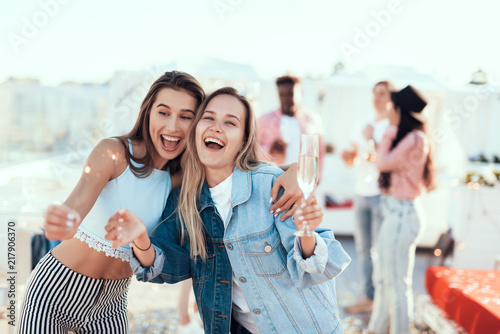 Portrait of optimistic lady and beaming girl holding firework sparklers in arms during party. Friends communicating behind them