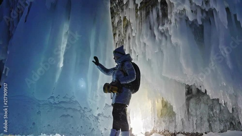 Travel woman on ice cave of Lake Baikal. Trip to winter island. Girl backpacker is walking of ice grot. Traveler looks at beautiful ice grotto. Hiker wears sports glasses, silver jacket, backpack, led