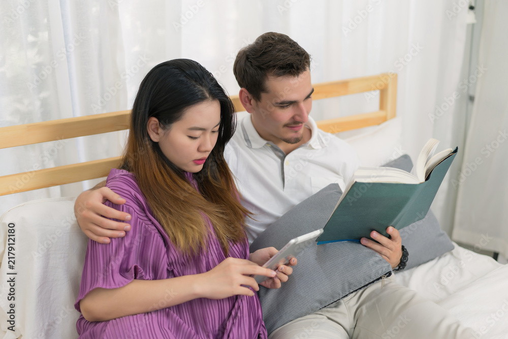Beautiful pregnant woman holding smart tablet computer and husband reading book on the bed in bedroom. Concept of couple, lover, family happy, lifestyle, relax and holiday. Beautiful pregnant woman holding smart tablet computer and husband reading book on the bed in bedroom. Concept of couple, lover, family happy, lifestyle, relax and holiday.