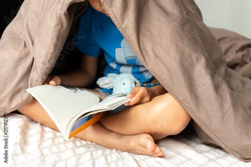 Young boy reading a book under the blanket in bed.