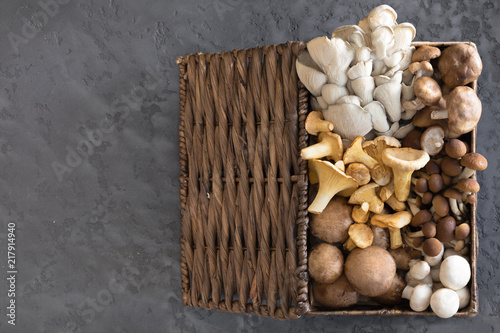 Top view of variety of uncooked wild forest mushrooms in a wicker basket on a black background, flat lay. Mushrooms chanterelles, honey agarics, oyster mushrooms, champignons, portobello, shiitake
