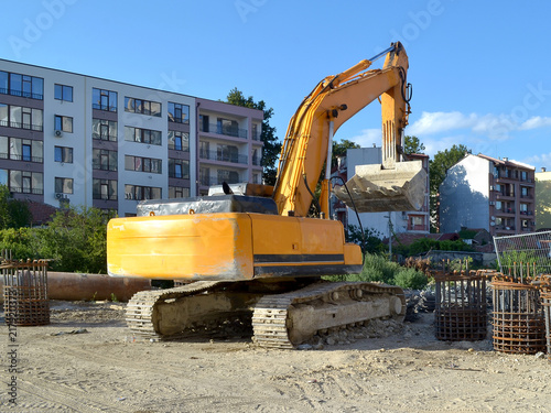 Yellow excavator at the site of road construction work near residential buildings, summer day, back view