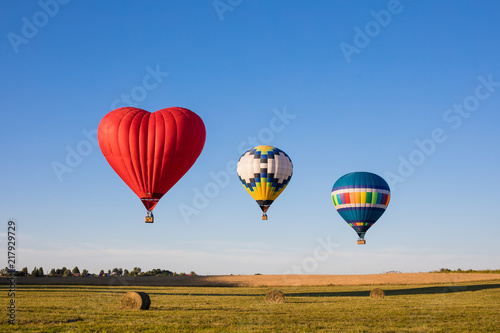 Red heart shaped and colorful air balloons flying over the fields with haystacks