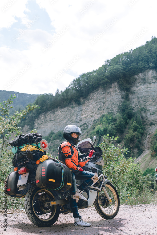 A girl dressed in body armor and helmet stand alone with big adventure touring motorcycle with bags and camping equipment, off road travel jorney, traveling together. mountain dirt road vertical photo A girl dressed in body armor and helmet stand alone with big adventure touring motorcycle with bags and camping equipment, off road travel jorney, traveling together. mountain dirt road vertical photo