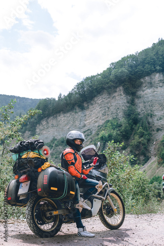 A girl dressed in body armor and helmet stand alone with big adventure touring motorcycle with bags and camping equipment  off road travel jorney  traveling together. mountain dirt road vertical photo