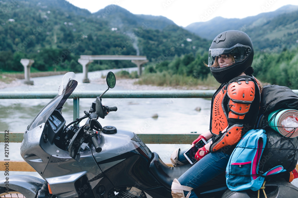 A girl in body armor and helmet sit on a big adventure touring motorcycle with bags and camping equipment, off road travel jorney, traveling together, couple, bridge, river mountains background