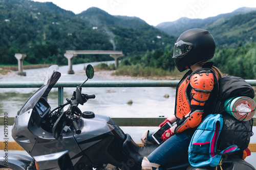 A girl in body armor and helmet sit on a big adventure touring motorcycle with bags and camping equipment, off road travel jorney, traveling together, couple, bridge, river mountains background
