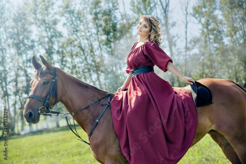 Beautiful girl in a red long red dress and in a black hat with a cocked hat riding a brown horse.