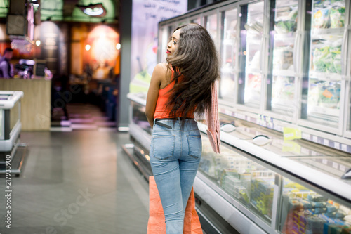 Pretty smiling african american girl in casual clothes holding pink paper bags in the supermarket. Shopping idea and concept photo