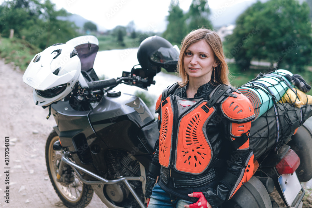 Fototapeta premium A girl dressed in body armor stand alone a with big adventure touring motorcycle with bags and camping equipment, off road travel jorney, traveling together, couple, mountains dirt road and river