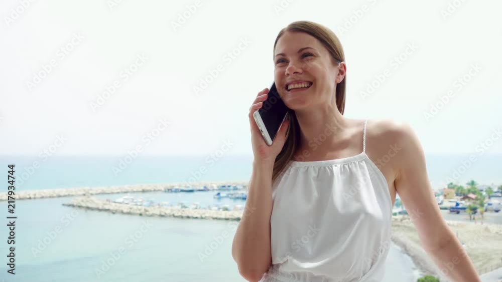 Smiling young woman in white dress using mobile on balcony. Female on vacation enjoying sea view from terrace in morning and talking to friend via cellphone in slow motion Smiling young woman in white dress using mobile on balcony. Female on vacation enjoying sea view from terrace in morning and talking to friend via cellphone in slow motion
