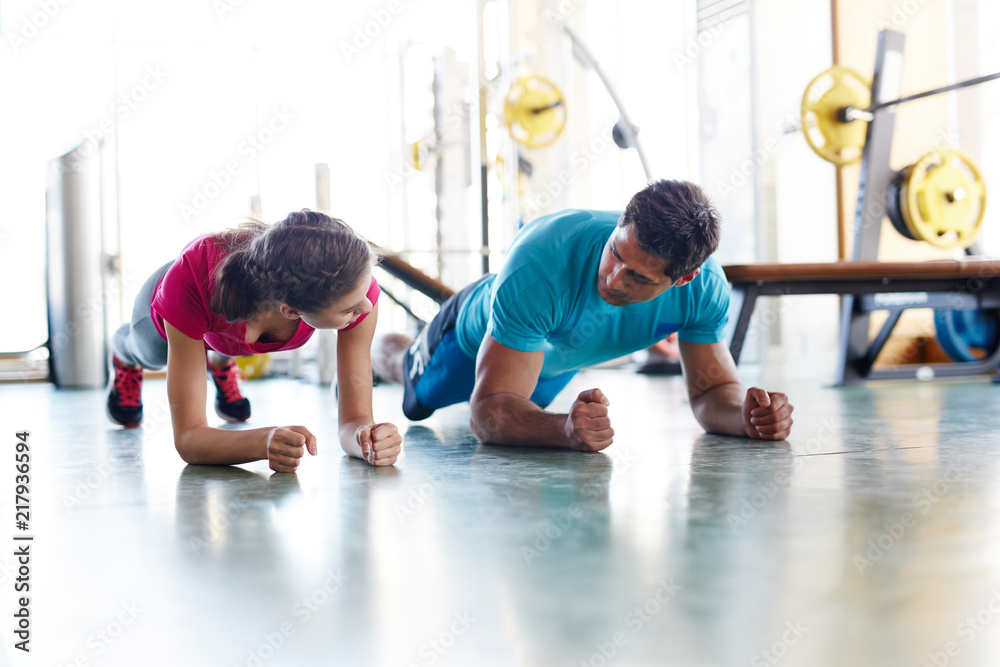Adult confident man and woman training together and doing plank exercise looking at each other in gym Adult confident man and woman training together and doing plank exercise looking at each other in gym