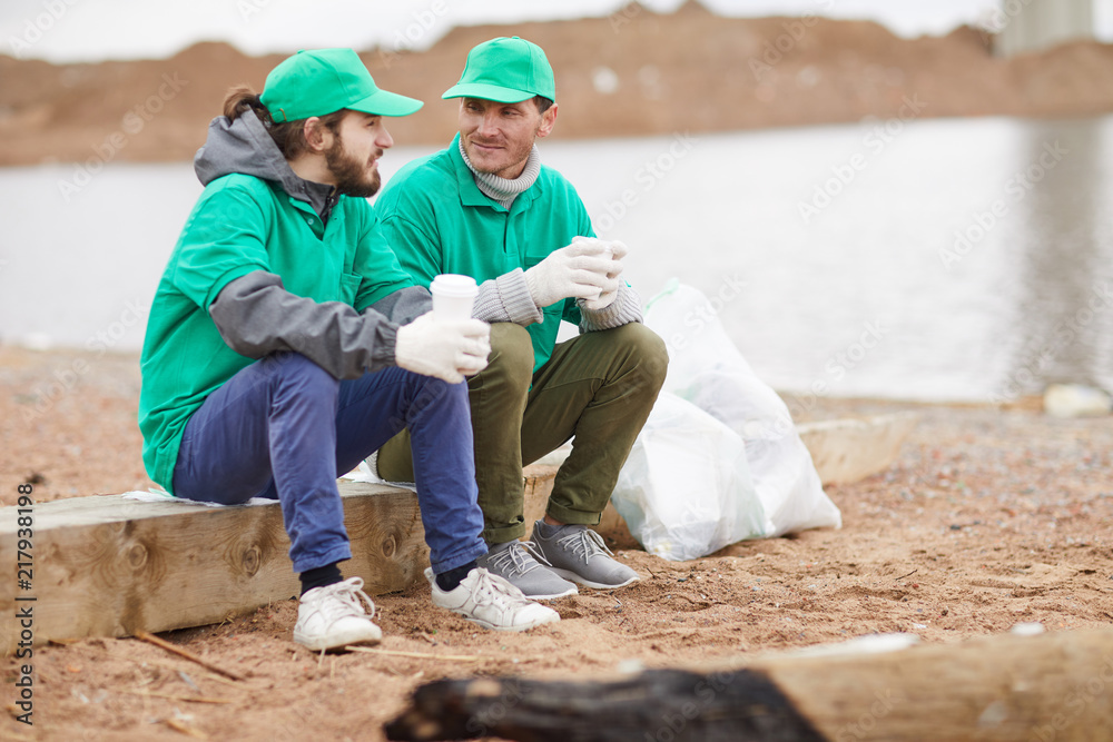 Two volunteers in green uniform sitting with paper cups and resting on polluted shore Two volunteers in green uniform sitting with paper cups and resting on polluted shore