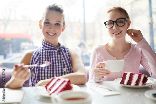 Cheerful young women sitting in cafe and eating delicious Red Velvet cake while looking at camera