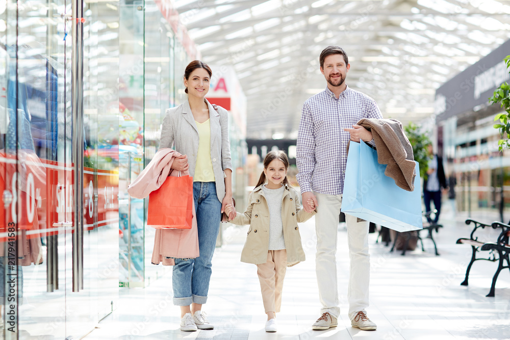 Obraz premium Adult cheerful parents with little girl carrying paper bags and standing in mall smiling at camera enjoying shopping together