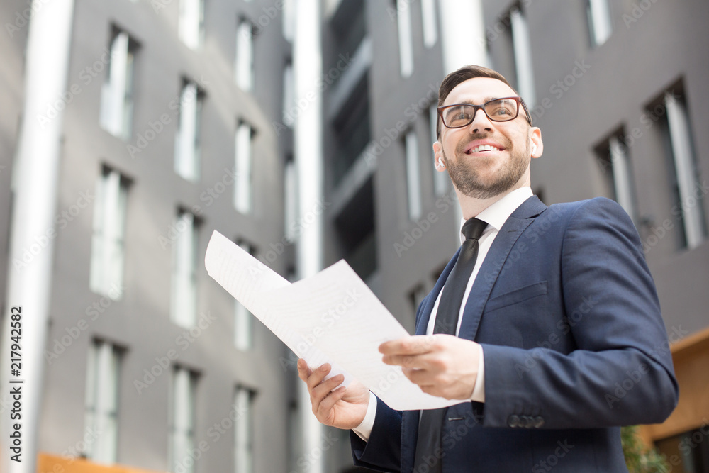From below shot of happy bearded man in suit and glasses holding papers and looking away on background of office building