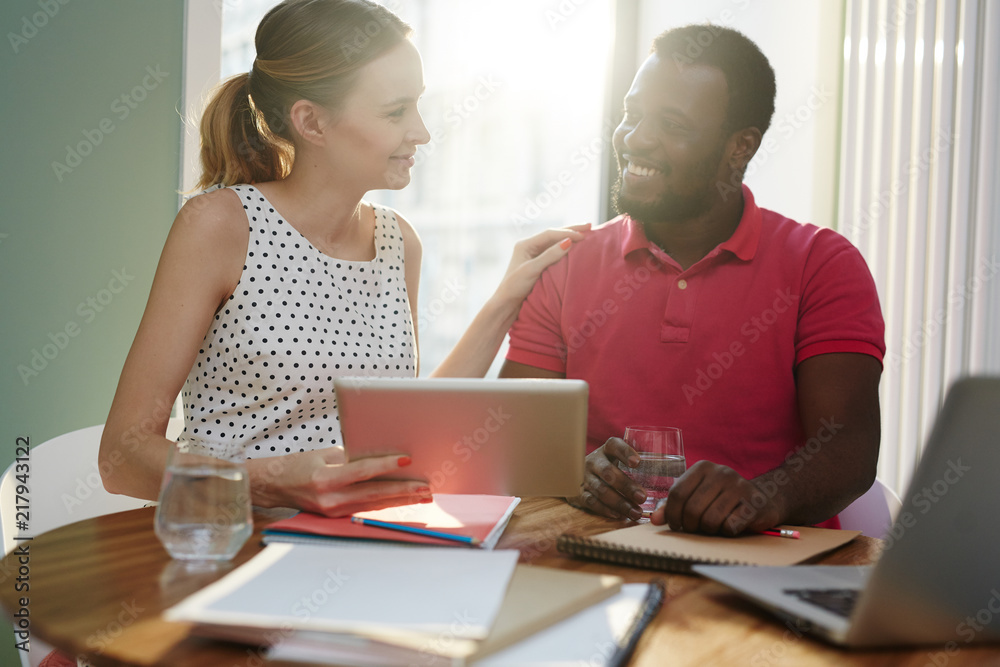 Smiling diverse man and woman sitting with tablet and notes at table and looking at each other working in team Smiling diverse man and woman sitting with tablet and notes at table and looking at each other working in team
