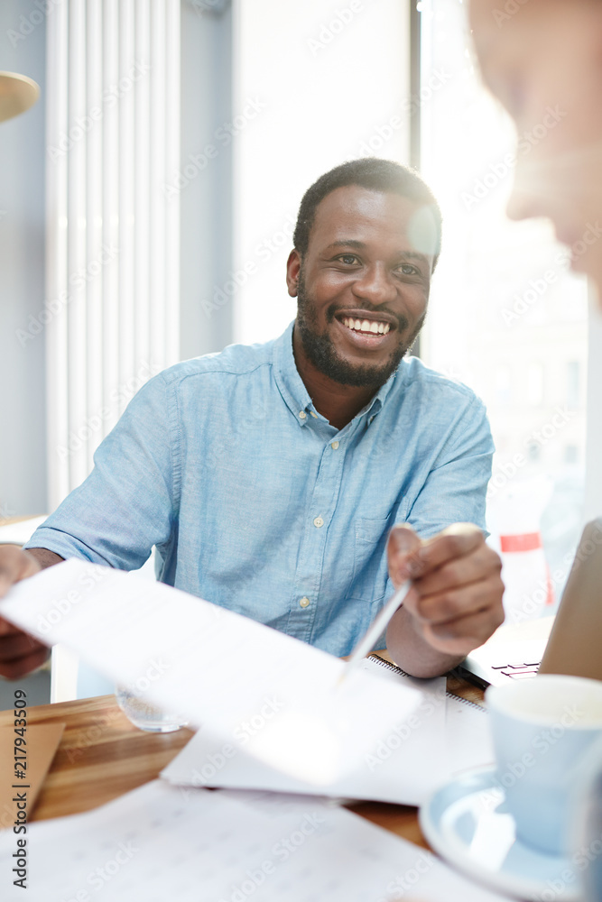 Handsome African-American man smiling and demonstrating documents to colleague while working in brightly lit office together