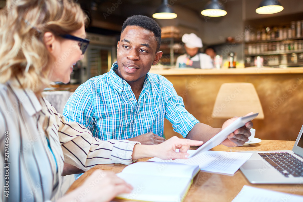 Side view of adult woman and black man smiling and browsing tablet together during business meeting in cafe Side view of adult woman and black man smiling and browsing tablet together during business meeting in cafe