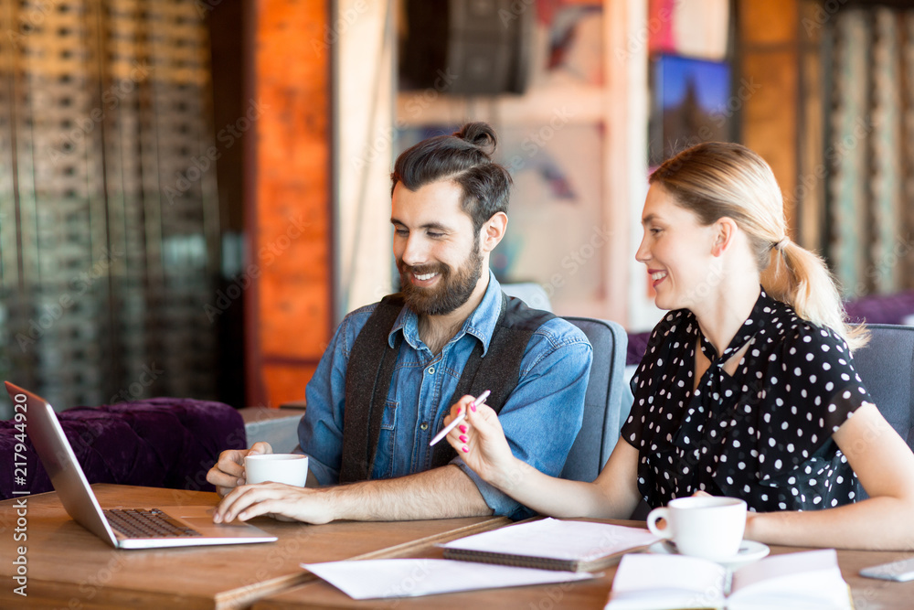 Trendy modern man and woman sitting at table in cafeteria watching laptop and having coffee