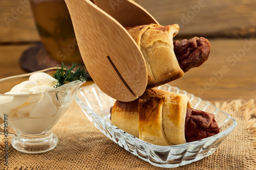 Baked puff pastry rolls stuffed with meat sausages in a glass plate, wooden kitchen tongs, sour cream sauce with dill, linen cloth napkin, all on a wooden background.
