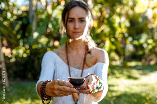 Oriental beauty. Closeup portrait of young beautiful woman holding out a cup of tea while seating against garden background