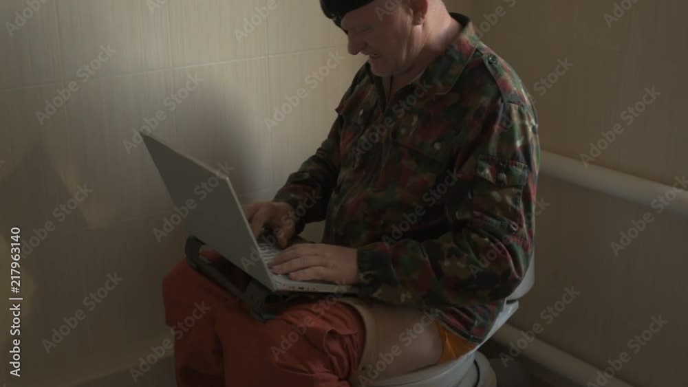 Man with gray hair, beard and mustache in military jacket with laptop sitting on toilet. Man's eye is covered with black medical patch. Dude is infatuated with computer and actively expressed emotions