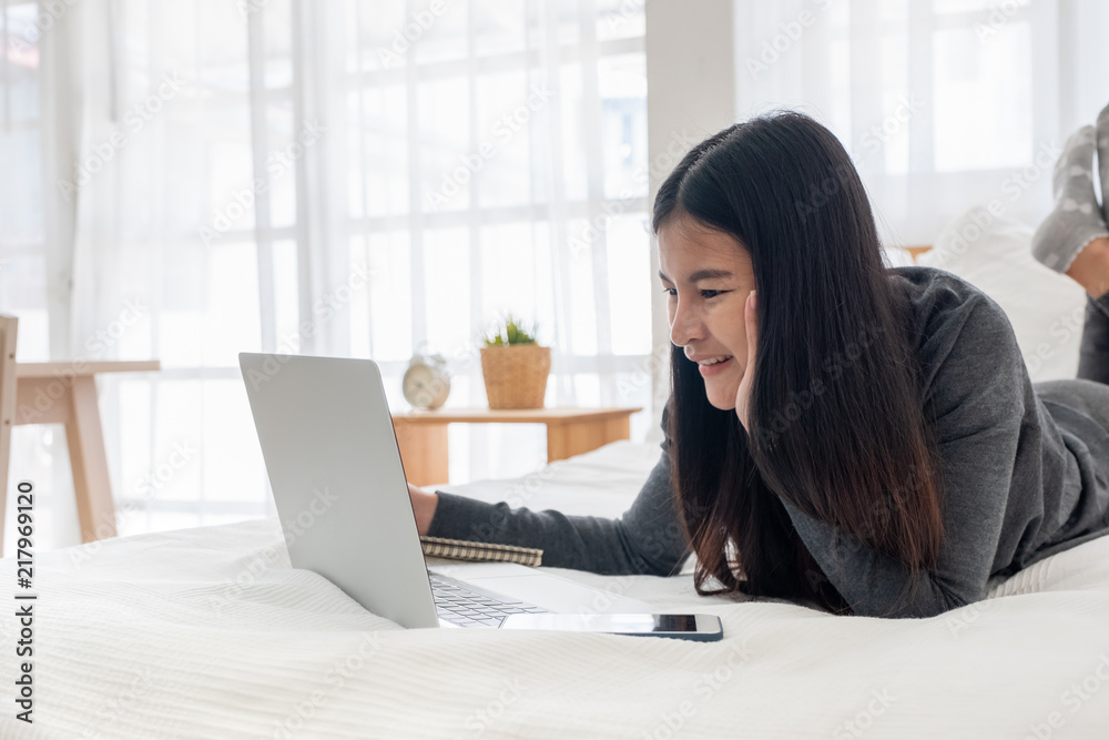 Asian woman laying on bed doing paperwork on laptop in bedroom.work at home.e-larning concept.study online. Asian woman laying on bed doing paperwork on laptop in bedroom.work at home.e-larning concept.study online.